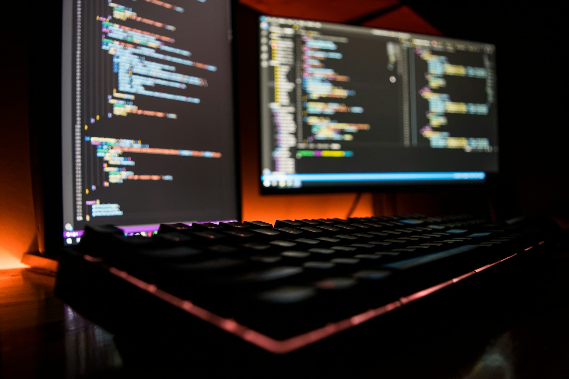 A keyboard glowing orange in front of two screens of code in a dark room